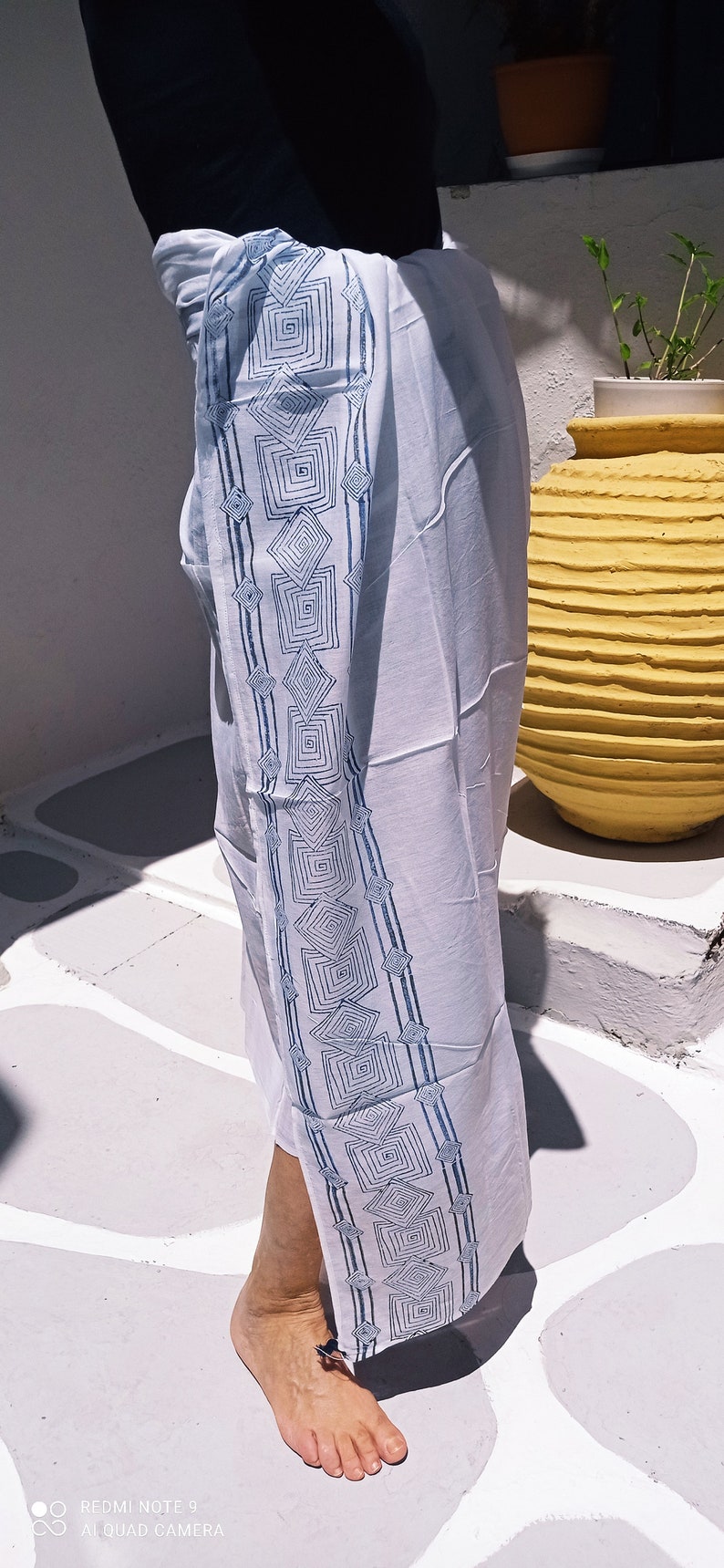 May include: White cotton sarong with a blue geometric pattern. The sarong is tied around the waist and has a long, flowing skirt.