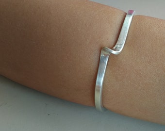 Puede incluir: Un brazalete de plata con un diseño curvo único. El brazalete se lleva en el brazo de una persona.