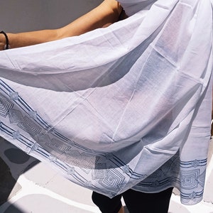 May include: A woman wearing a white cotton scarf with a blue geometric pattern. The scarf is draped over her shoulders and she is standing on a white tiled floor.