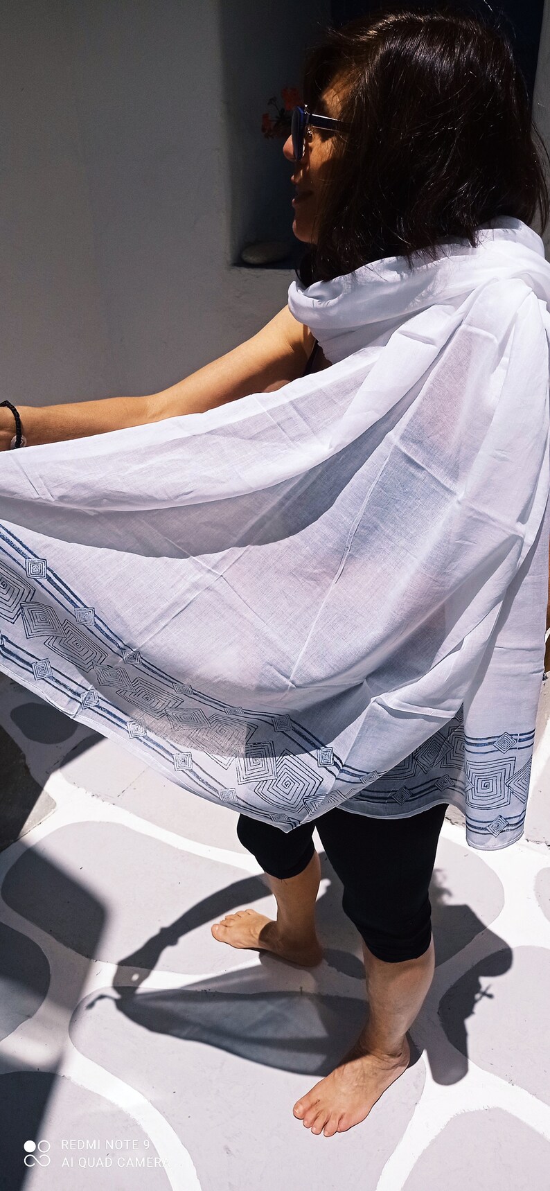 May include: A woman wearing a white cotton scarf with a blue geometric pattern. The scarf is draped over her shoulders and she is standing on a white tiled floor.