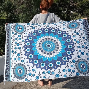 May include: A white tapestry with a blue and white mandala design featuring an evil eye motif. The tapestry is hanging from a person's hands.