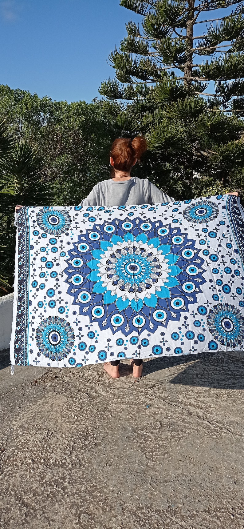 May include: A white tapestry with a blue and white mandala design featuring an evil eye motif. The tapestry is hanging from a person's hands.