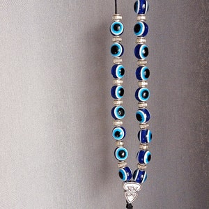 May include: A necklace with blue and white evil eye beads. The beads are strung on a black cord with silver-coloured metal accents. The necklace is displayed against a grey background.