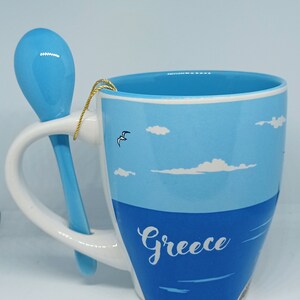 May include: A ceramic mug with a blue and white design, featuring the word "Greece" and a matching blue spoon. The mug has a handle and a design of the sea and sky.