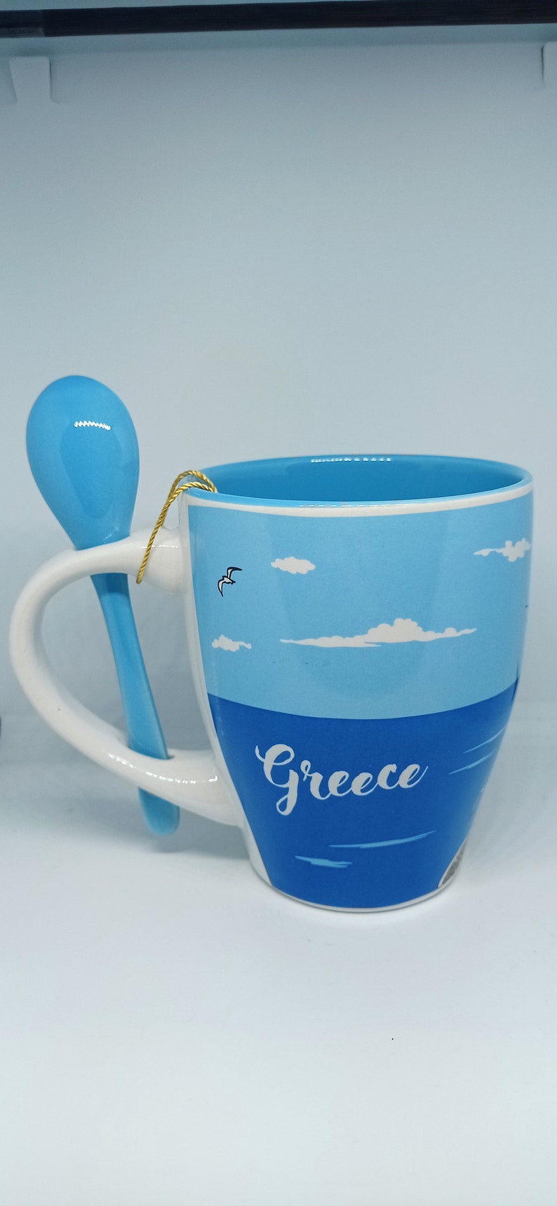 May include: A ceramic mug with a blue and white design, featuring the word "Greece" and a matching blue spoon. The mug has a handle and a design of the sea and sky.