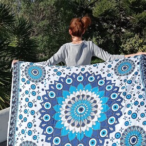 May include: A white tapestry with a blue and white mandala design featuring an evil eye motif. The tapestry is held up by a person in front of a green tree.
