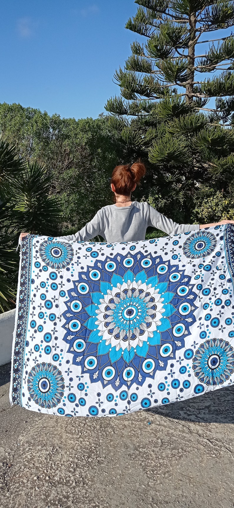 May include: A white tapestry with a blue and white mandala design featuring an evil eye motif. The tapestry is held up by a person in front of a green tree.