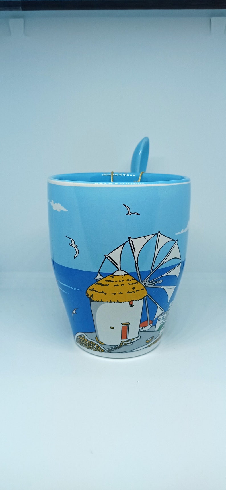 May include: A light blue ceramic mug with a matching spoon. The mug features a seaside illustration with a windmill, seagulls, and a blue sky. The spoon rests on the rim, held by a gold wire.