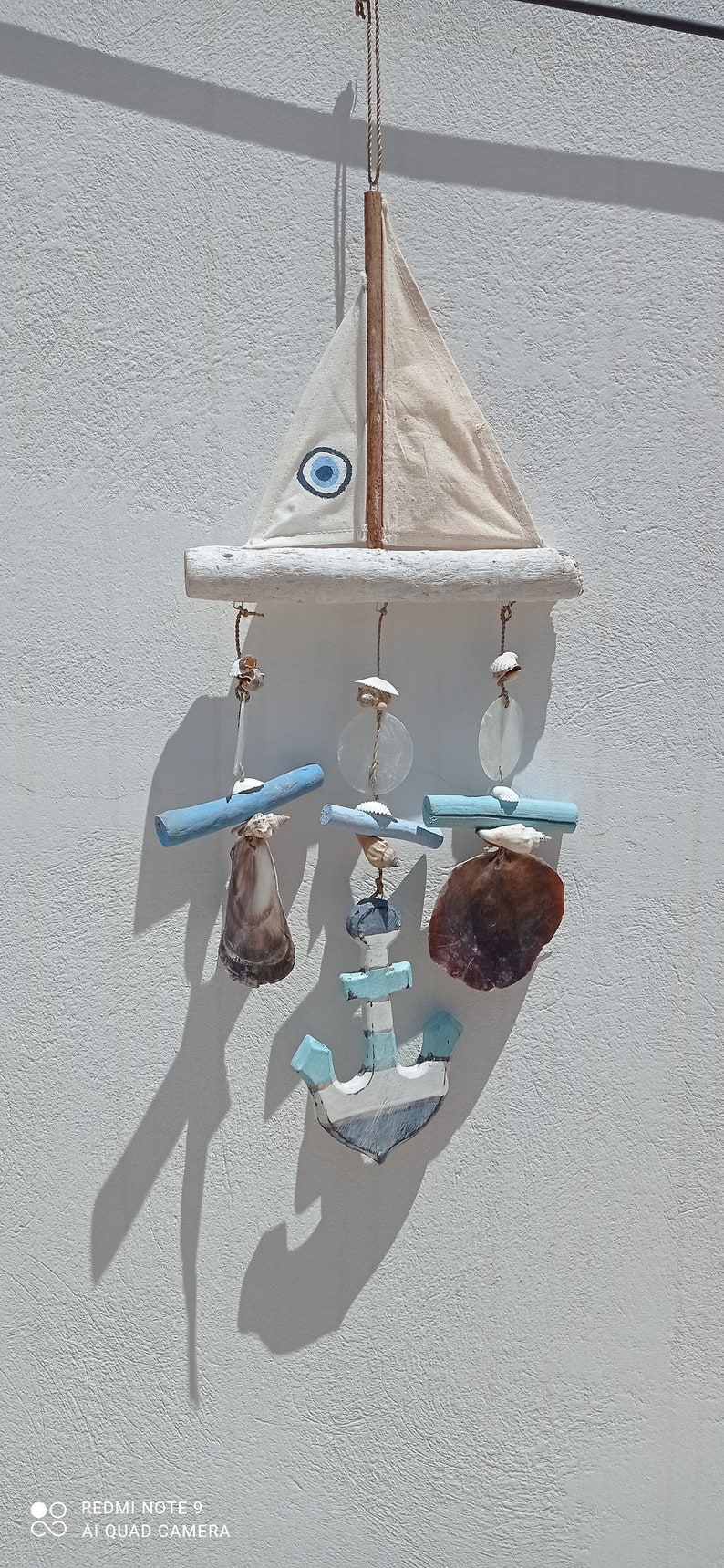 Może przedstawiać: Biały żaglowiec z niebieskim amuletem w kształcie złego oka wiszący na gałęzi dryfującego drewna. Żaglowiec jest ozdobiony muszelkami i niebiesko-białą kotwicą.