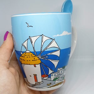 May include: A ceramic mug with a scenic illustration of a Greek island windmill and village. The mug is light blue with a matching spoon resting on the rim. The design includes a windmill, sea, and buildings.