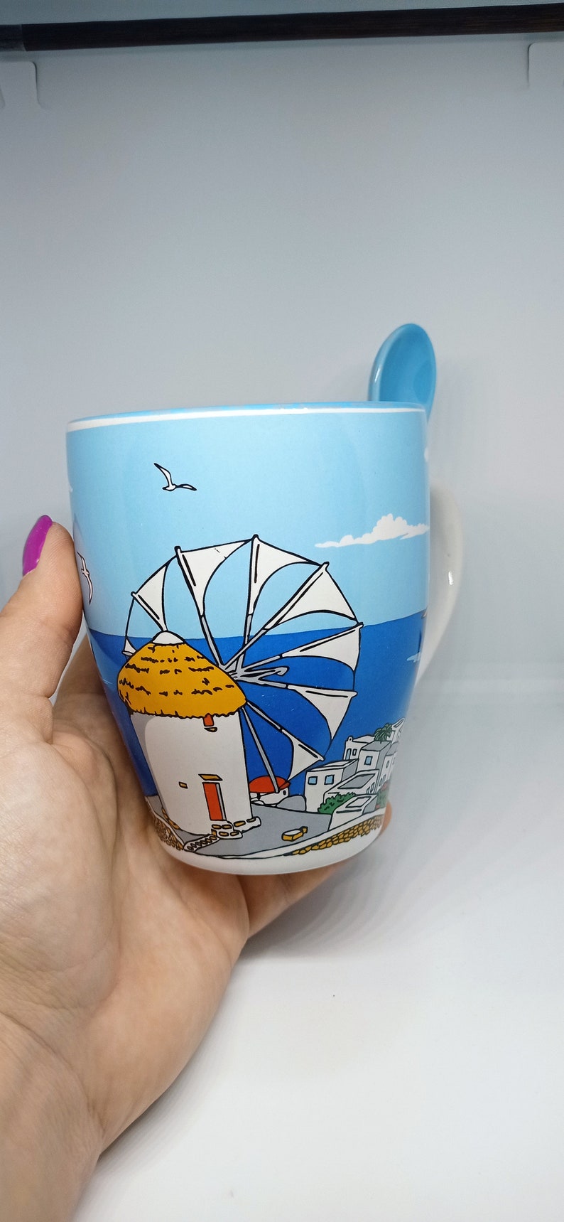 May include: A ceramic mug with a scenic illustration of a Greek island windmill and village. The mug is light blue with a matching spoon resting on the rim. The design includes a windmill, sea, and buildings.