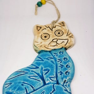 May include: A ceramic cat ornament with a blue and white glaze. The cat has a whimsical expression and is decorated with a floral design. The ornament is strung with a twine cord and has two small wooden beads.