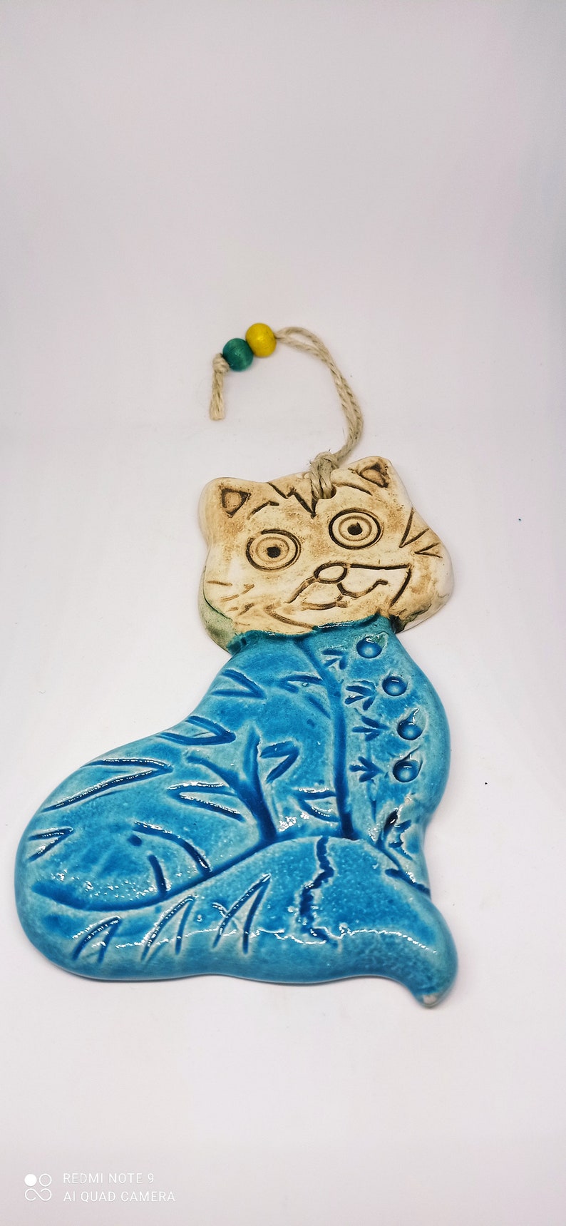 May include: A ceramic cat ornament with a blue and white glaze. The cat has a whimsical expression and is decorated with a floral design. The ornament is strung with a twine cord and has two small wooden beads.