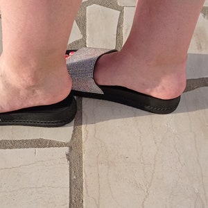 May include: A pair of black slide sandals with a sparkly, rhinestone strap. The sandals are on a light-coloured stone floor.