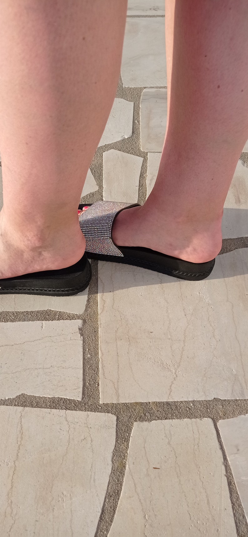 May include: A pair of black slide sandals with a sparkly, rhinestone strap. The sandals are on a light-coloured stone floor.