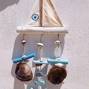 Może przedstawiać: Biały drewniany żaglowiec z niebieskim amuletem złego oka wiszący na białej ścianie. Żaglowiec ma niebieską i białą kotwicę wiszącą z niego, a z kotwicy zwisają trzy muszle.