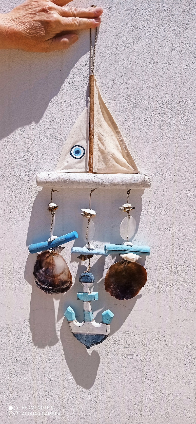 Może przedstawiać: Biały drewniany żaglowiec z niebieskim amuletem złego oka wiszący na białej ścianie. Żaglowiec ma niebieską i białą kotwicę wiszącą z niego, a z kotwicy zwisają trzy muszle.
