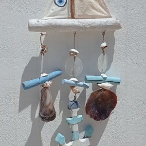 Może przedstawiać: Biały żaglowiec z niebieskim amuletem w kształcie złego oka wisi na gałęzi dryfującego drewna. Żaglowiec jest ozdobiony niebieskimi i białymi muszelkami oraz niebieską i białą kotwicą.