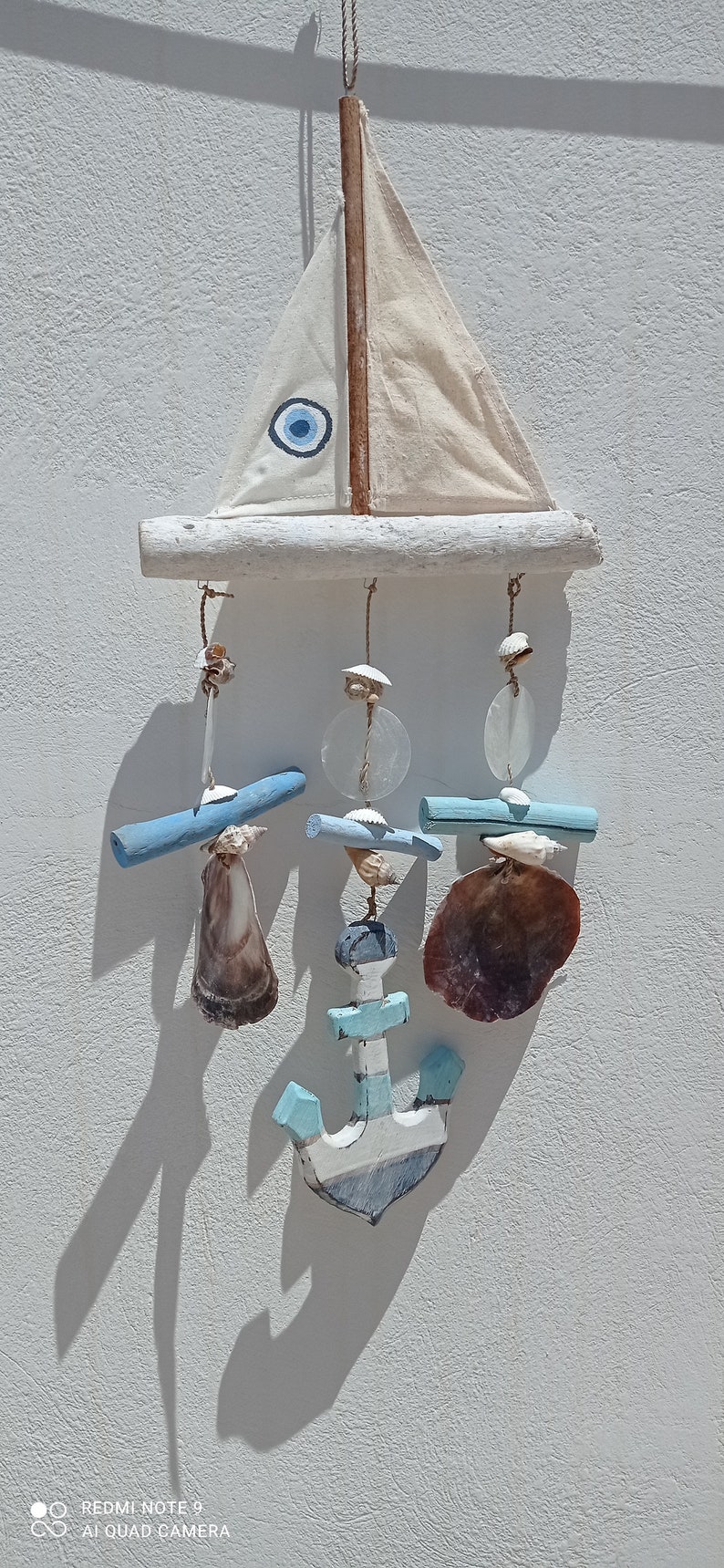 Może przedstawiać: Biały żaglowiec z niebieskim amuletem w kształcie złego oka wisi na gałęzi dryfującego drewna. Żaglowiec jest ozdobiony niebieskimi i białymi muszelkami oraz niebieską i białą kotwicą.