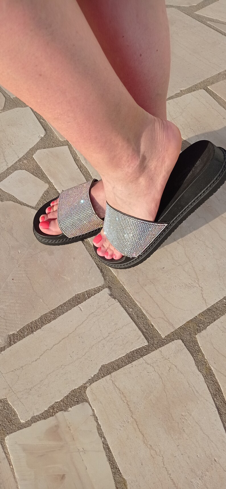 May include: A pair of black slide sandals with a sparkly silver strap. The sandals have a thick black sole and are decorated with rhinestones.