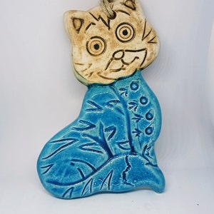 May include: A ceramic cat figurine with a blue glaze. The cat is sitting with its head tilted and has a whimsical expression. The figurine is hand-painted with details on the cat's face and body.