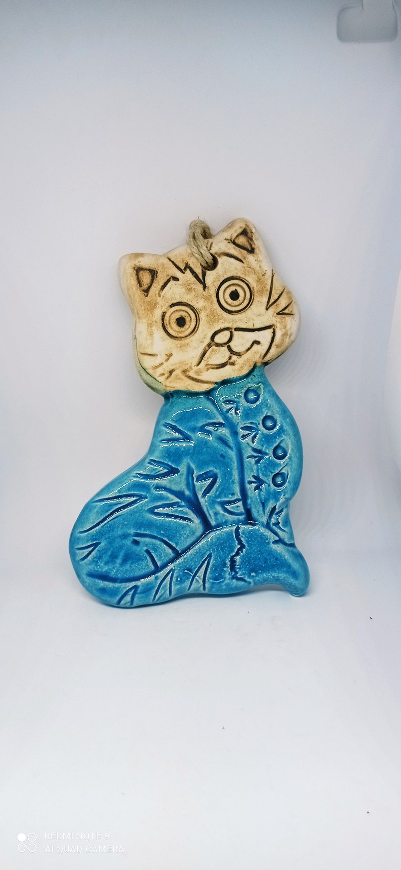 May include: A ceramic cat figurine with a blue glaze. The cat is sitting with its head tilted and has a whimsical expression. The figurine is hand-painted with details on the cat's face and body.