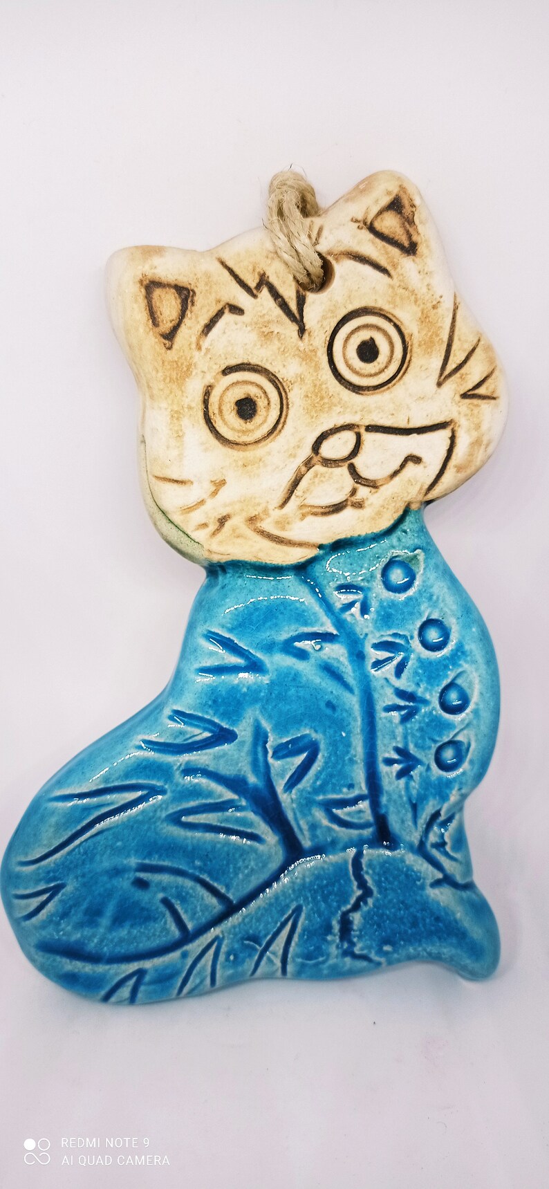 May include: A ceramic cat ornament with a blue and white glaze. The cat has a white face with brown eyes and a brown nose. The cat is wearing a blue coat with a floral pattern.