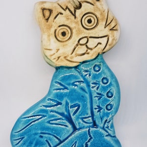 May include: A ceramic cat ornament with a blue and white glaze. The cat has a white face with brown eyes and a brown mouth. The cat is sitting on a blue background with white leaves.