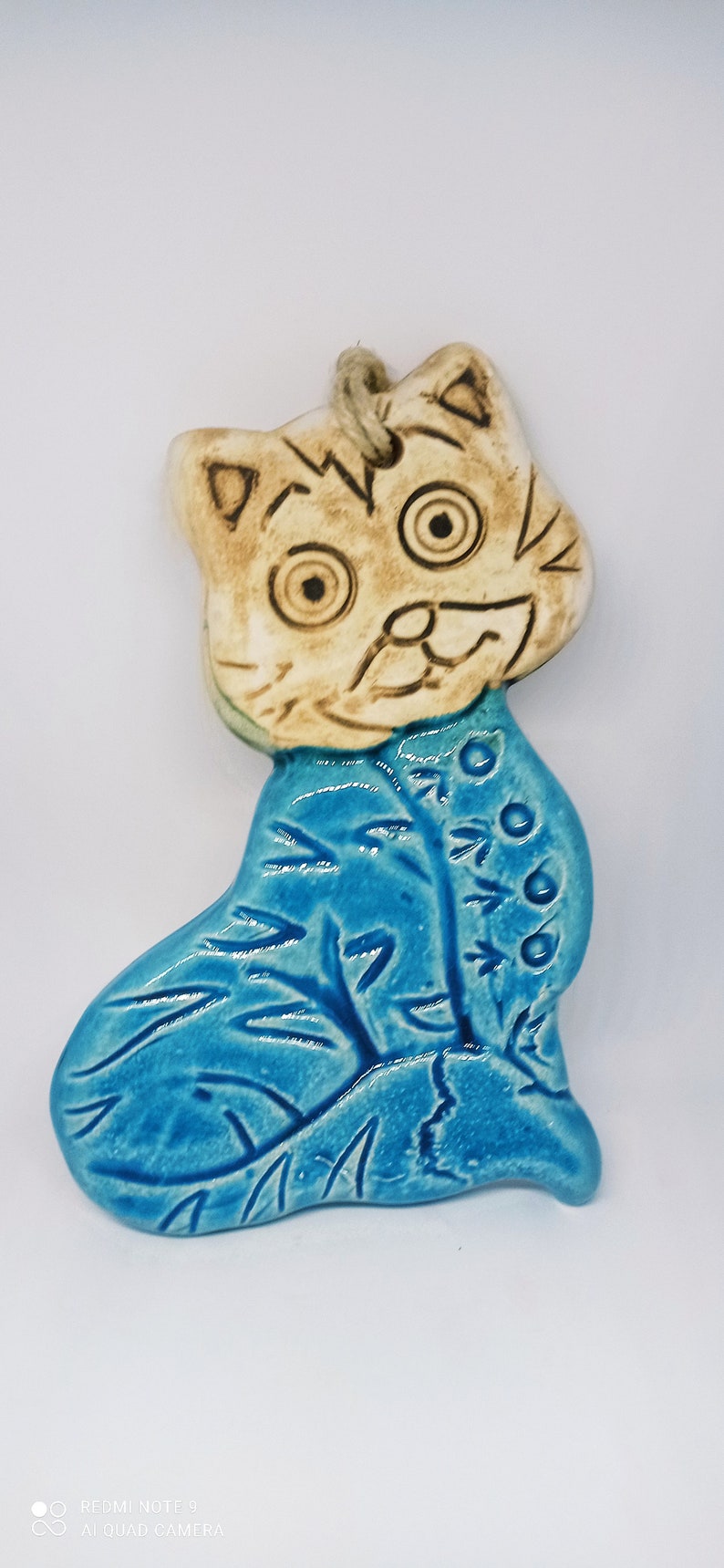 May include: A ceramic cat ornament with a blue and white glaze. The cat has a white face with brown eyes and a brown mouth. The cat is sitting on a blue background with white leaves.