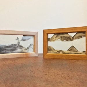 Può includere: Due cornici per l'arte della sabbia, una con una cornice in legno chiaro e l'altra con una cornice in legno scuro. Entrambe le cornici contengono arte della sabbia con un design paesaggistico.