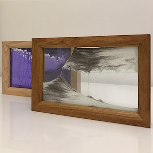 Peut inclure: Deux cadres en bois contenant des œuvres d'art de sable. La plus grande pièce présente un motif de sable gris avec un motif ondulé. La plus petite pièce présente un motif de sable violet avec un motif tourbillonnant.