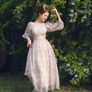 May include: A light beige dress with a blue floral pattern. The dress has long, sheer sleeves and a flowing skirt. The person in the image is wearing a floral crown and standing in a garden setting.
