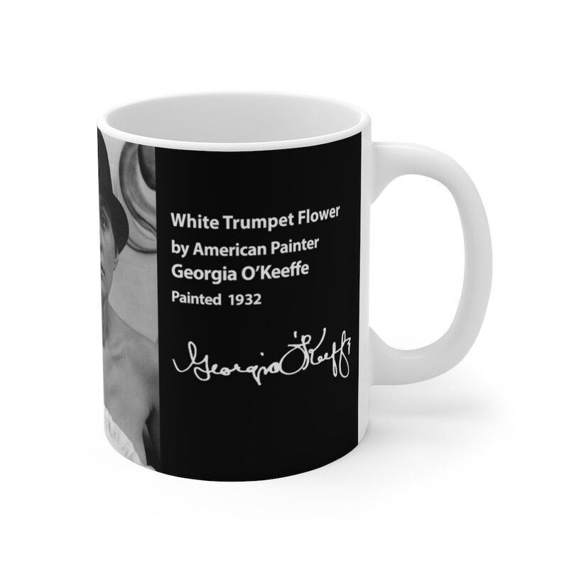 Op de afbeelding: Witte keramische mok met een zwart-witte afbeelding van een vrouw en tekst die luidt "White Trumpet Flower by American Painter Georgia O'Keeffe Painted 1932" en een handtekening die "Georgia O'Keeffe" luidt.
