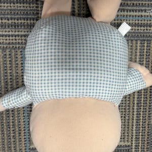 May include: A plush bunny toy, viewed from the back, features a light pink body and ears. The bunny's back and arms are covered in a blue and white gingham pattern. The toy is lying on a patterned carpet.