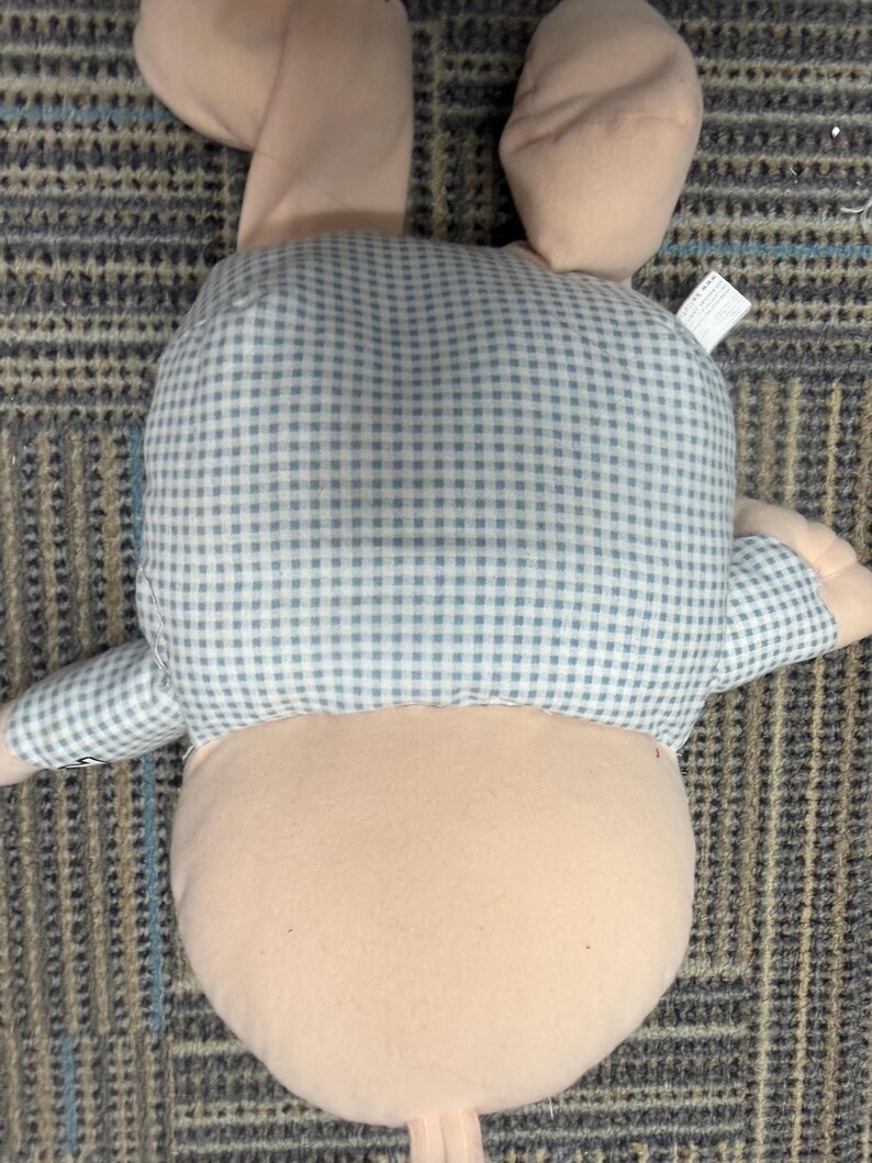 May include: A plush bunny toy, viewed from the back, features a light pink body and ears. The bunny's back and arms are covered in a blue and white gingham pattern. The toy is lying on a patterned carpet.