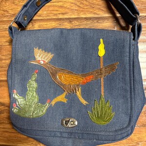 May include: A blue denim shoulder bag featuring an embroidered roadrunner bird, a green cactus with red accents, and a tall yellow cattail plant. The bag has a flap closure secured with a silver-toned metal twist lock and a matching denim strap.