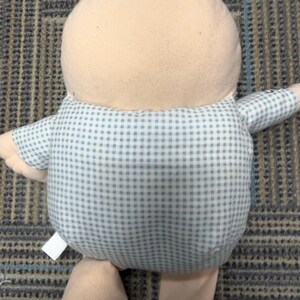 May include: A plush doll with a light tan head and limbs, and a blue and white checkered shirt. The doll has a rounded body and is positioned upright. The doll's fabric appears soft and cuddly, suggesting it is a children's toy.