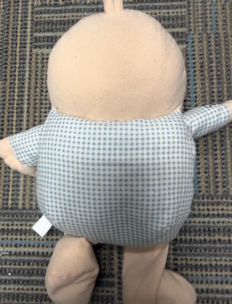 May include: A plush doll with a light tan head and limbs, and a blue and white checkered shirt. The doll has a rounded body and is positioned upright. The doll's fabric appears soft and cuddly, suggesting it is a children's toy.