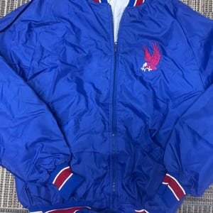 May include: A blue nylon bomber jacket with a red and white striped collar, cuffs, and waistband. The jacket features a red and pink embroidered eagle emblem on the chest. The jacket has a zipper closure.