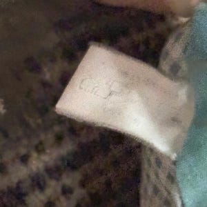 May include: A close-up of a white tag with the text "C.J." in a cursive font. The tag is slightly crumpled and appears to be attached to a soft, textured fabric. The background is blurred, with hints of blue and patterned fabric.