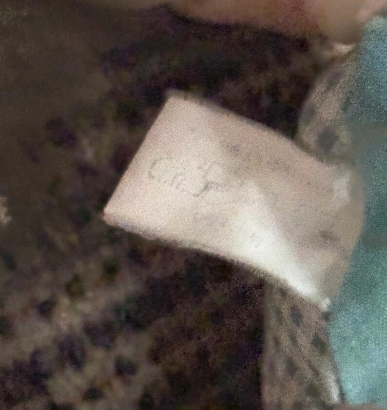 May include: A close-up of a white tag with the text "C.J." in a cursive font. The tag is slightly crumpled and appears to be attached to a soft, textured fabric. The background is blurred, with hints of blue and patterned fabric.