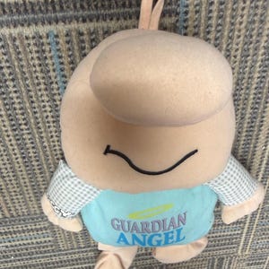 May include: A plush toy in the shape of a smiling figure with a large head and small arms and legs. The toy is primarily beige with a light blue shirt that reads "GUARDIAN ANGEL" in blue text. The arms are checkered.
