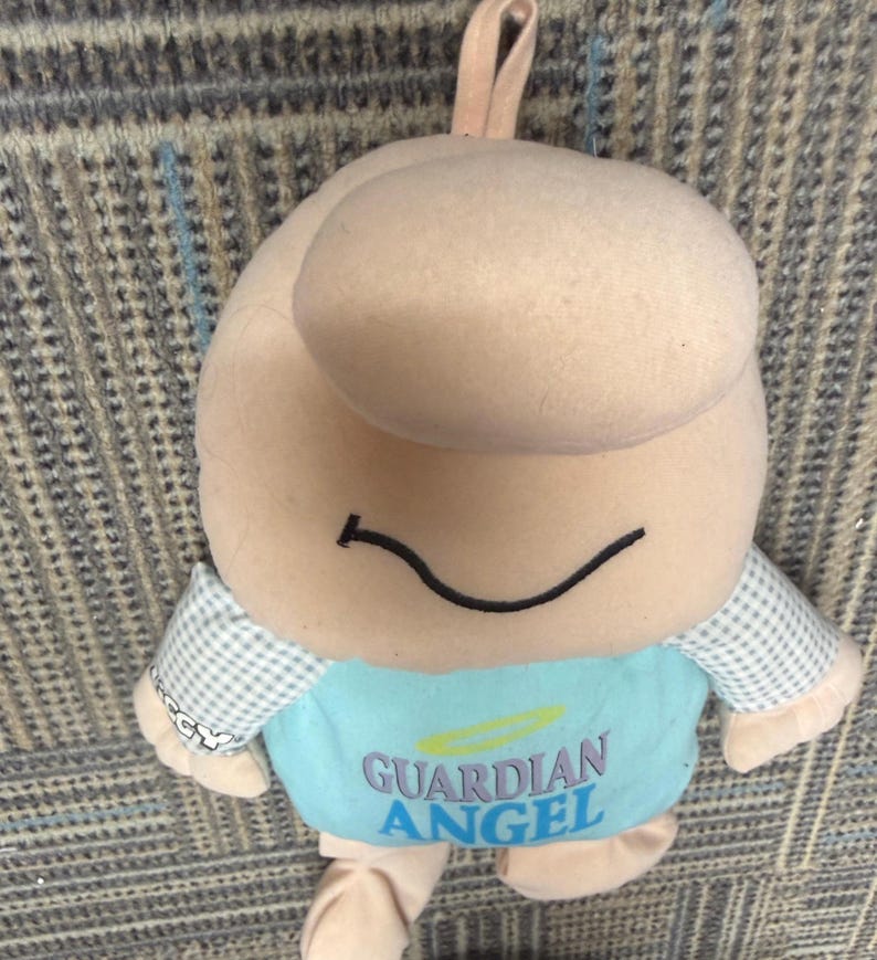 May include: A plush toy in the shape of a smiling figure with a large head and small arms and legs. The toy is primarily beige with a light blue shirt that reads "GUARDIAN ANGEL" in blue text. The arms are checkered.