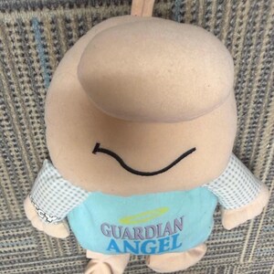 May include: A plush toy in the shape of a smiling figure with a large head and small arms and legs. The figure is primarily beige with a blue shirt that reads "GUARDIAN ANGEL". The sleeves are a light blue and white checkered pattern.