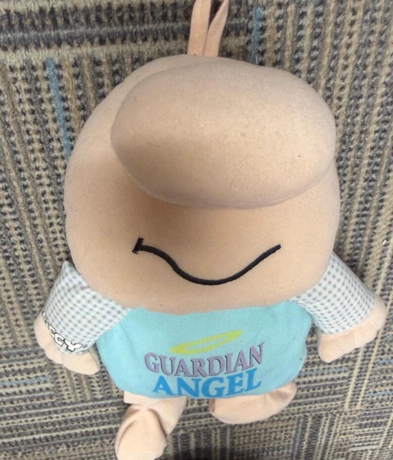 May include: A plush toy in the shape of a smiling figure with a large head and small arms and legs. The figure is primarily beige with a blue shirt that reads "GUARDIAN ANGEL". The sleeves are a light blue and white checkered pattern.