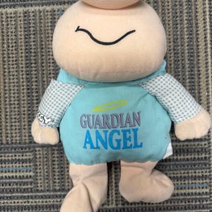 May include: A plush toy in the shape of a smiling figure with a light pink head and body. It has a blue shirt with the words "GUARDIAN ANGEL" printed on it. The arms and feet are light pink. The sleeves are checkered.
