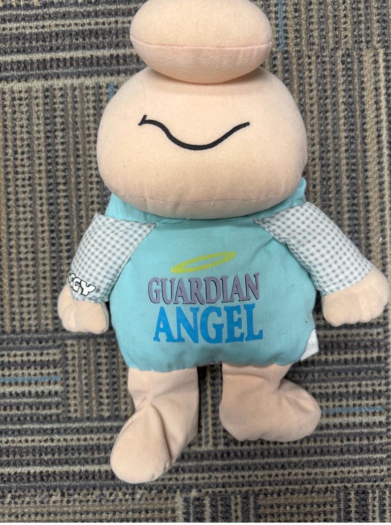May include: A plush toy in the shape of a smiling figure with a light pink head and body. It has a blue shirt with the words "GUARDIAN ANGEL" printed on it. The arms and feet are light pink. The sleeves are checkered.