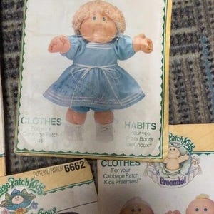 May include: Vintage Cabbage Patch Kids pattern packet featuring a doll in a blue dress. The packet includes the text "CLOTHES" and "HABITS" in English and French, with the Cabbage Patch Kids logo.