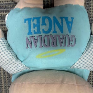 May include: A plush doll with a light blue diaper featuring the words "GUARDIAN ANGEL" in blue and purple. The doll has light pink skin and checkered blue and white wings.
