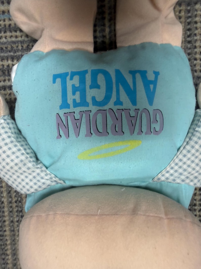 May include: A plush doll with a light blue diaper featuring the words "GUARDIAN ANGEL" in blue and purple. The doll has light pink skin and checkered blue and white wings.